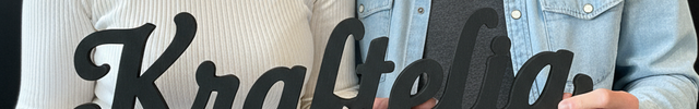 Zwei Personen sitzen nebeneinander und halten ein Holzschild mit der Aufschrift ‚Kraftélia‘ in den Händen.